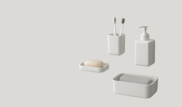 A collection of modern, minimalist white bathroom accessories including a soap dish with a bar of soap, a toothbrush holder with two toothbrushes, a soap dispenser, and a small tray with a neatly rolled grey towel.