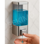 A hand dispensing liquid soap from a wall-mounted chrome and transparent dispenser.